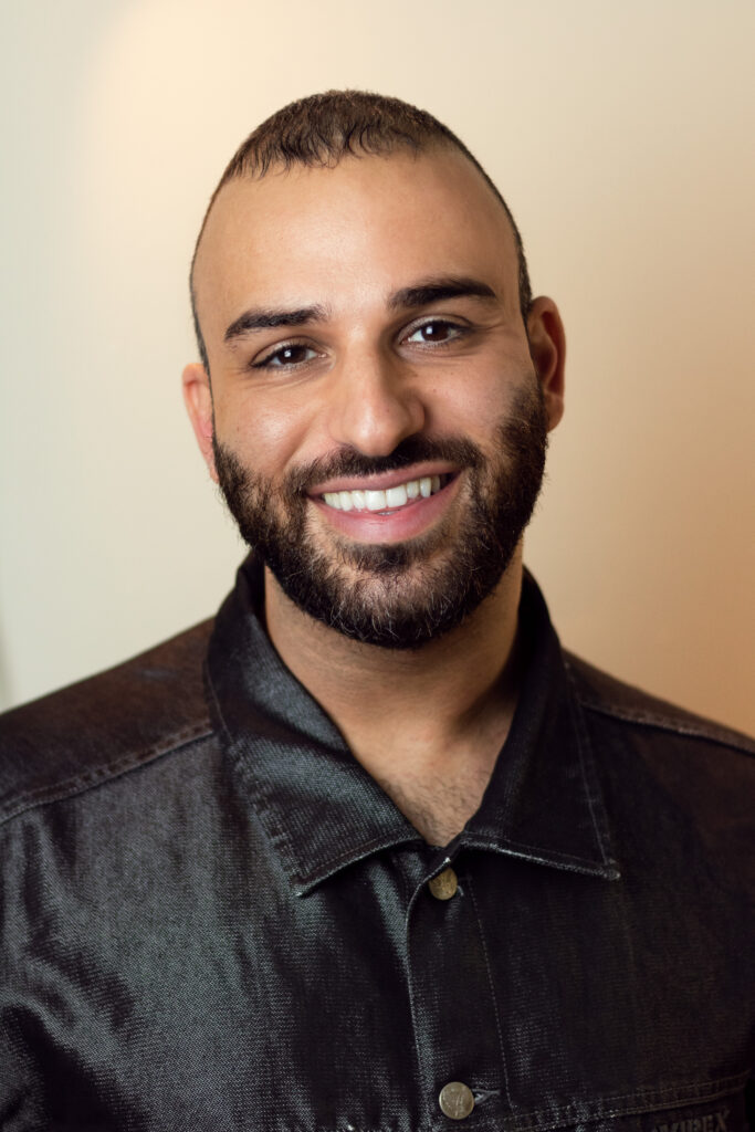 A headshot of a person with a beard, wearing a black shiny shirt, smiling at the camera